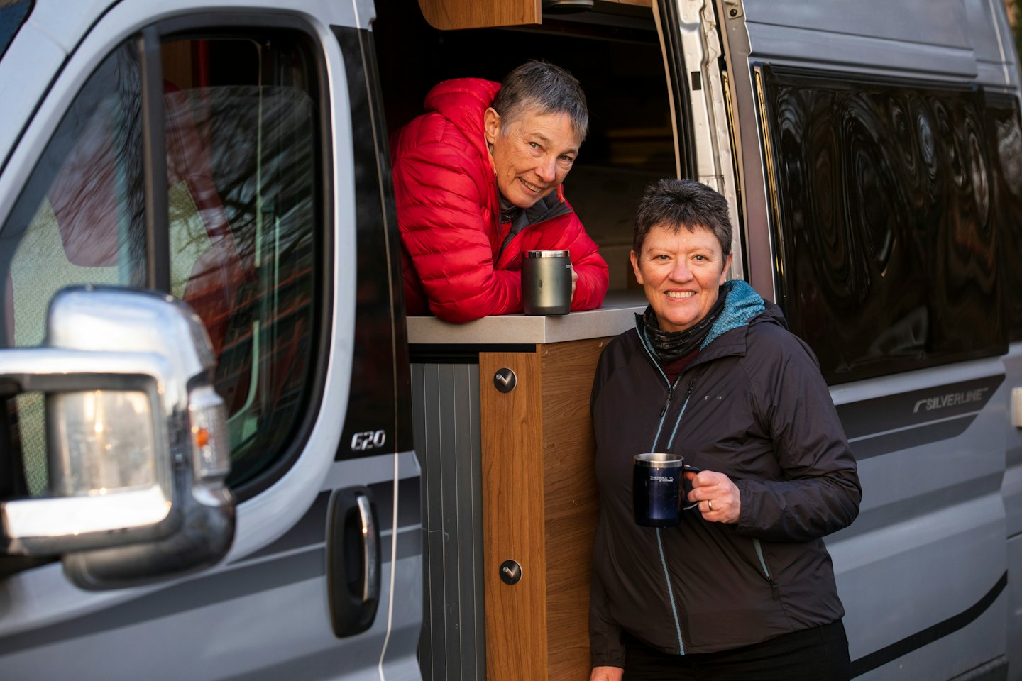 Two women holding travel mugs standing by a camper van.